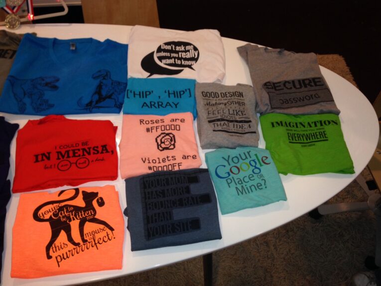 A display of various colorful graphic t-shirts laid out on a white surface, each featuring different humorous and nerdy text and designs by The PaperStreet Team.