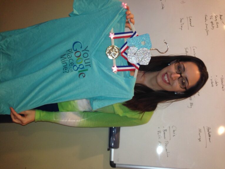 A woman wearing glasses and a green top holds up a blue shirt with "Your Google Place or Mine?" written on it, a T-shirt design by the PaperStreet Team. She also displays several medals. A whiteboard with writing is visible in the background.