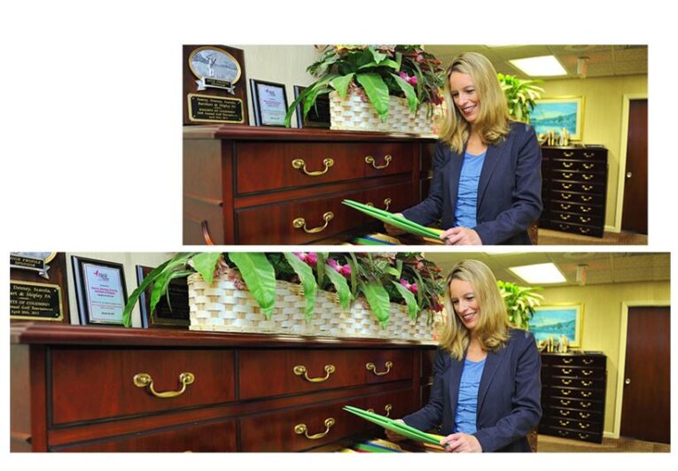 A woman stands next to a wooden cabinet holding green folders, smiling as she looks at the documents. Various plaques and awards are displayed on the cabinet, and plants adorn the top. An open book titled "Part 2: Using Photoshop to Enhance Your Website Photographs" lies nearby, reflecting her interest in digital creativity.