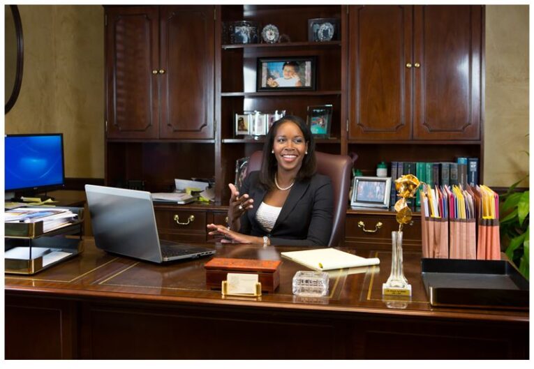 A woman sits at an office desk with a laptop, smiling while holding a pen. The desk is organized with files, a phone, and stationery. Wooden cabinets and shelves with books and photos are in the background as she reviews "Part 2: Using Photoshop to Enhance Your Website Photographs.