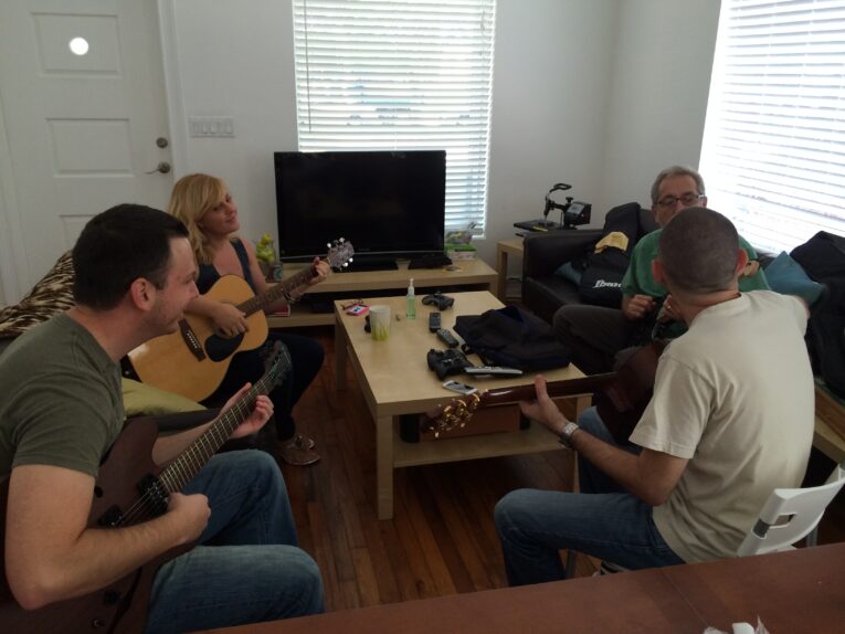 Four people are sitting in a living room playing guitars, with two men and two women. A television, table, and various items are in the background—a perfect scene depicting Guitar Lessons at PaperSteet.