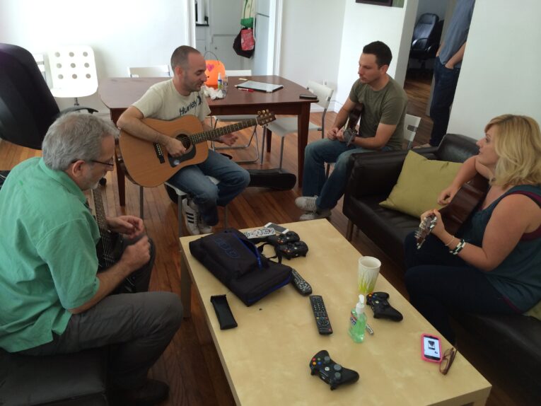 Four people are sitting in a living room playing guitars and chatting, likely here for Guitar Lessons at PaperStreet. Game controllers and a tablet rest on the coffee table in the center.