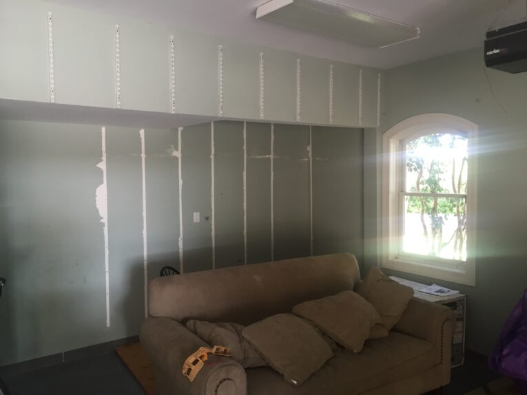 A beige couch with multiple cushions is placed against a wall in a room with unfinished drywall, reminiscent of those DIY spaces where you might learn how to build a rock climbing wall for kids in the garage. A window on the right side lets in natural light.