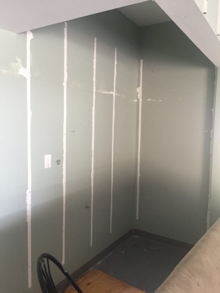 A small, unfinished room with gray walls featuring white vertical seams and a bare floor. There's a metal chair partially visible in the corner, making it seem like an ideal space for those wondering how to build a rock climbing wall for kids in the garage.