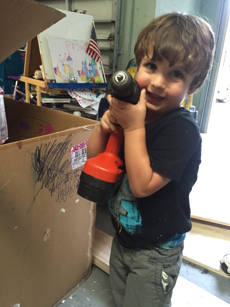 A young boy stands in a cluttered room holding an orange power drill. Behind him, a cardboard box with scribbles on it and an easel with a partially painted picture are visible—a scene reminiscent of a "How to Build a Rock Climbing Wall for Kids in the Garage" tutorial come to life.