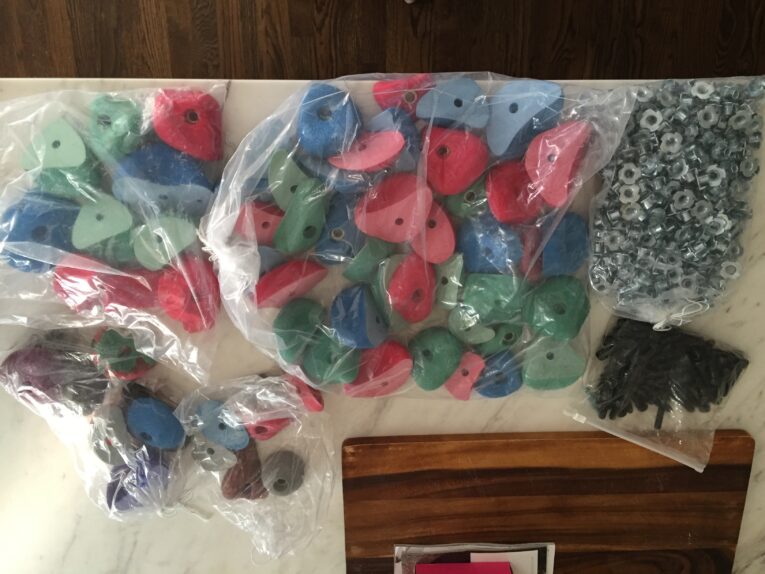 Bags of colorful rock climbing holds, metal hardware, and black screws are spread out on a white table, ready for the next step in learning how to build a rock climbing wall for kids in the garage.