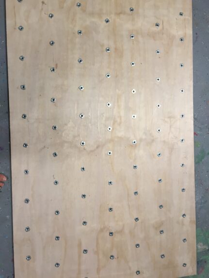A wooden board with evenly spaced metal nuts embedded in it, displayed flat on a surface, is essential for learning how to build a rock climbing wall for kids in the garage.