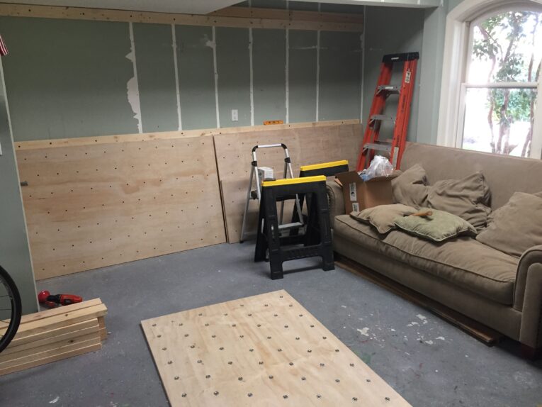 A room under renovation with plywood sheets, saw horses, a ladder, and a sofa with cushions. The wall is partially stripped, exposing studs—perfect for those considering how to build a rock climbing wall for kids in the garage.