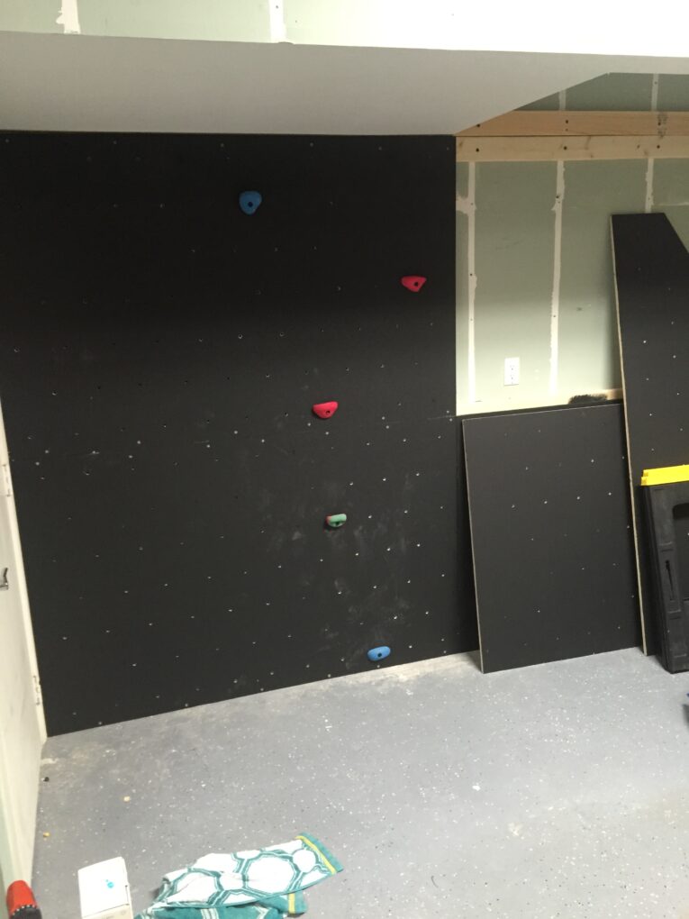 A partially constructed indoor climbing wall with five visible handholds stands in a room with unfinished walls and gray flooring, offering the first steps in understanding how to build a rock climbing wall for kids in the garage.