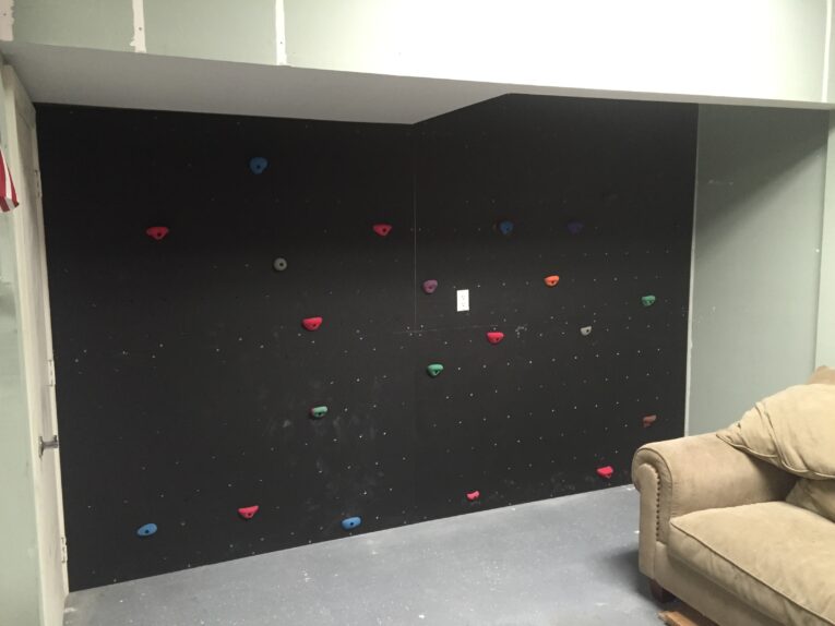 An indoor climbing wall with colorful handholds, resembling those found in guides on how to build a rock climbing wall for kids in the garage, is installed on a black wall in a room with an unfinished ceiling. A beige sofa is positioned to the right of the climbing wall.