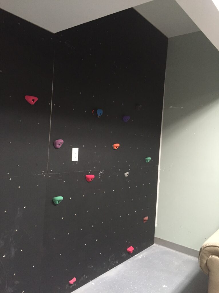 An indoor climbing wall with multi-colored holds is set against a black wall in a room with gray and beige walls, offering inspiration on how to build a rock climbing wall for kids in the garage.