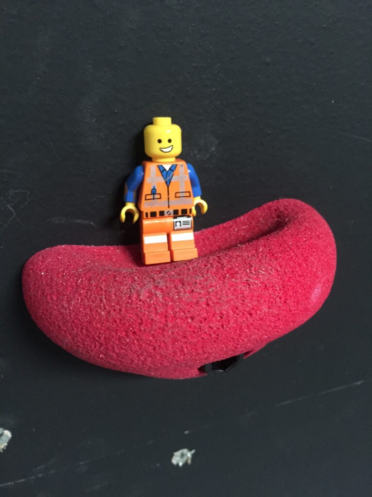 A LEGO figurine dressed as a construction worker is placed on a red climbing hold mounted on a black wall, demonstrating how to build a rock climbing wall for kids in the garage.