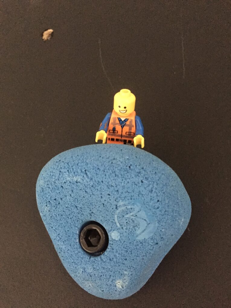 A Lego figurine is placed on top of a blue rock climbing hold with a bolt in the center, showcasing an example of how to build a rock climbing wall for kids in the garage. The hold is securely attached to a sturdy black climbing wall.