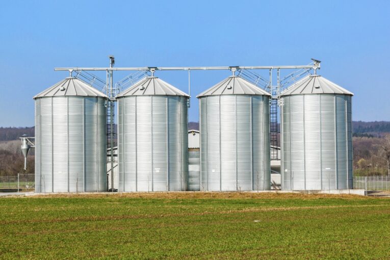 Four large silver grain silos stand in a grassy field under a clear blue sky, much like how the content on your website fills with valuable information, ready to harvest.