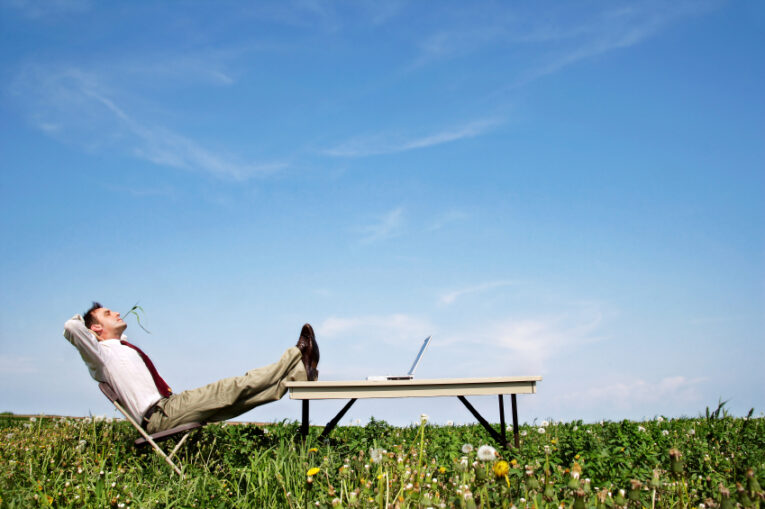 A man in a business attire relaxes on a chair in a grassy field, his legs propped on a table with a laptop, gazing at the sky.