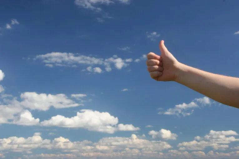 A hand giving a thumbs-up gesture is set against a backdrop of a blue sky with scattered white clouds.