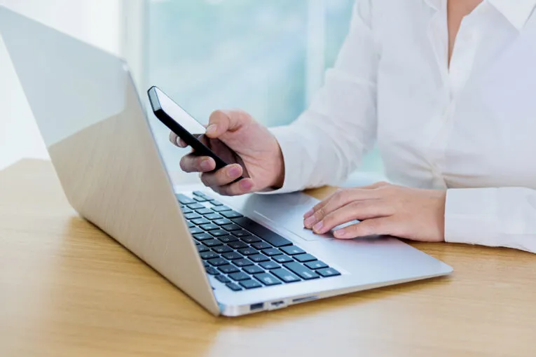 A person in a white shirt reads "Website Launch Tips - A Best Practices Guide" on their smartphone while working on a laptop at a desk.
