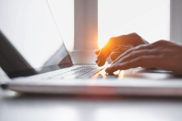 A person typing on a laptop in a bright room with sunlight streaming through a window.