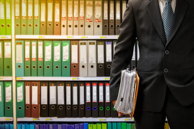 A person in a suit holding a stack of documents stands in front of shelves filled with colorful binders and folders.