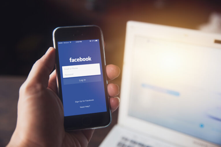 A hand holding a smartphone displaying the Facebook login screen, with a laptop open in the background.