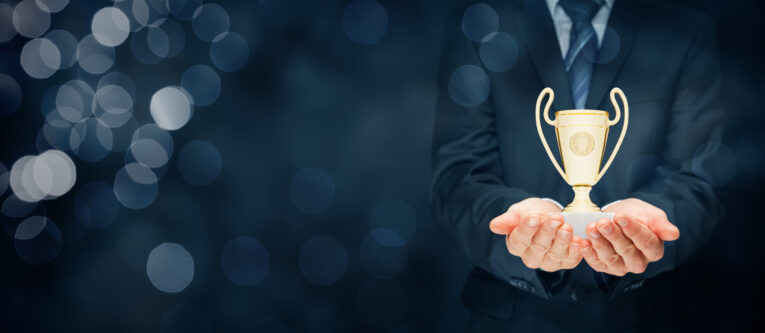 A person in a suit holds a small gold trophy with both hands, against a dark, bokeh-lit background.