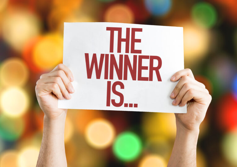 Two hands holding a sign with the text "The Winner Is..." against a colorful, blurred background suggest the anticipation and excitement of bidding on the competition's name.