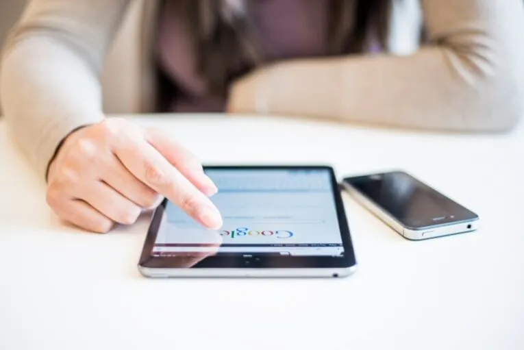 A person is using a tablet with the Google homepage visible on the screen. There is also a smartphone placed on the table beside the tablet.