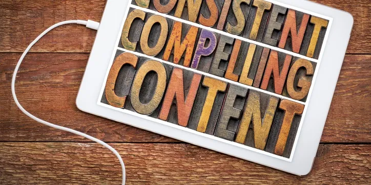 A tablet screen displaying the words "Consistent Compelling Content" in colorful block letters, with an attached white charging cable, all placed on a wooden surface.