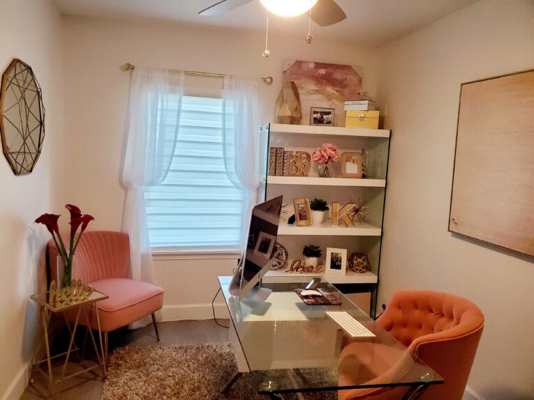 A small home office with a glass desk, computer, keyboard, mouse, pink chairs, a bookshelf with decorative items, a window with sheer curtains, and wall art.