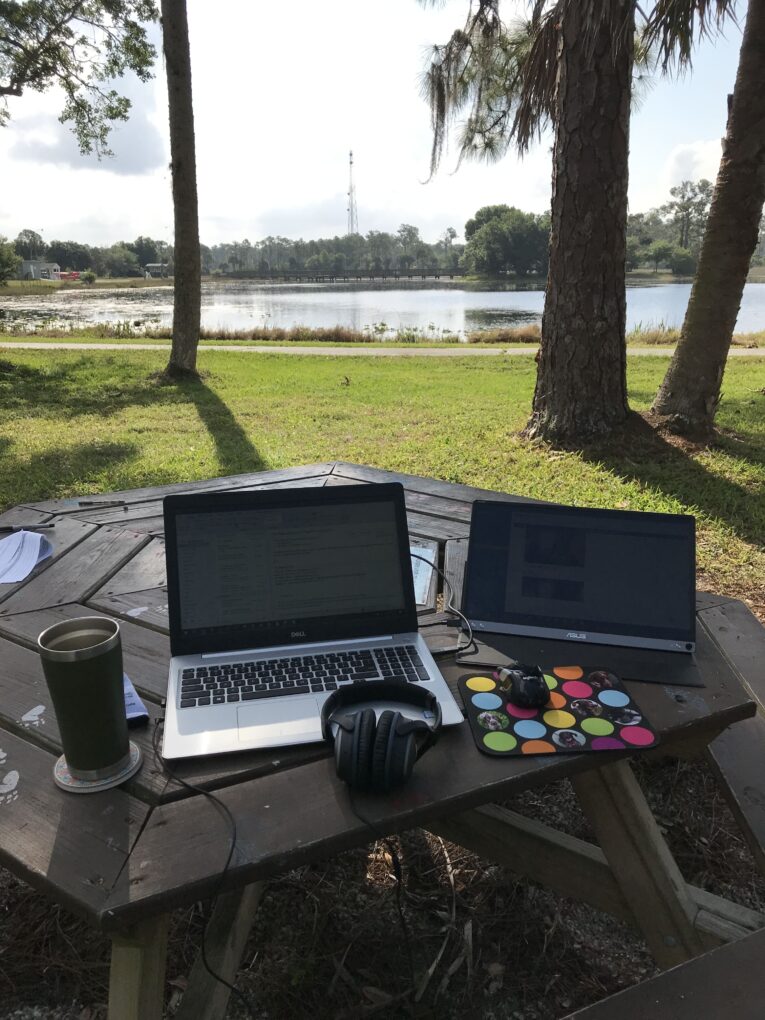 Outdoor setup with two laptops on a wooden picnic table, a pair of headphones, a colorful mouse pad, and a drink. The table is near a lake surrounded by trees.