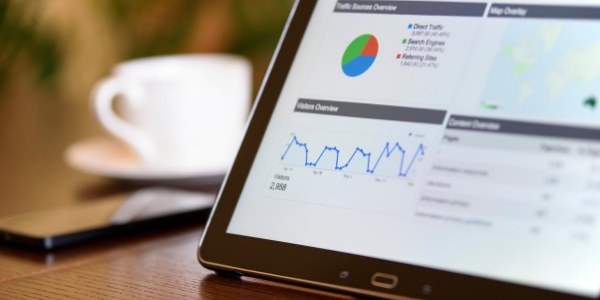 A tablet on a wooden table displays website analytics, including a pie chart and line graph detailing how to use Google Analytics to identify exit pages and improve them. A blurred coffee cup and smartphone are in the background.