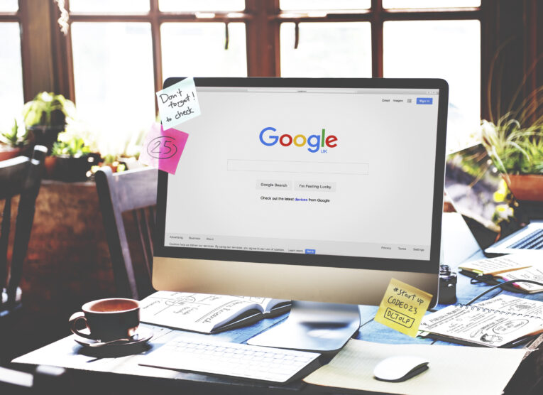 A computer on a desk displaying the Google UK homepage. The desk has a cup, papers, notebooks, and sticky notes. The background shows windows with sunlight coming in, providing an ideal setting to explore Local Services Ads - How to Get Started.