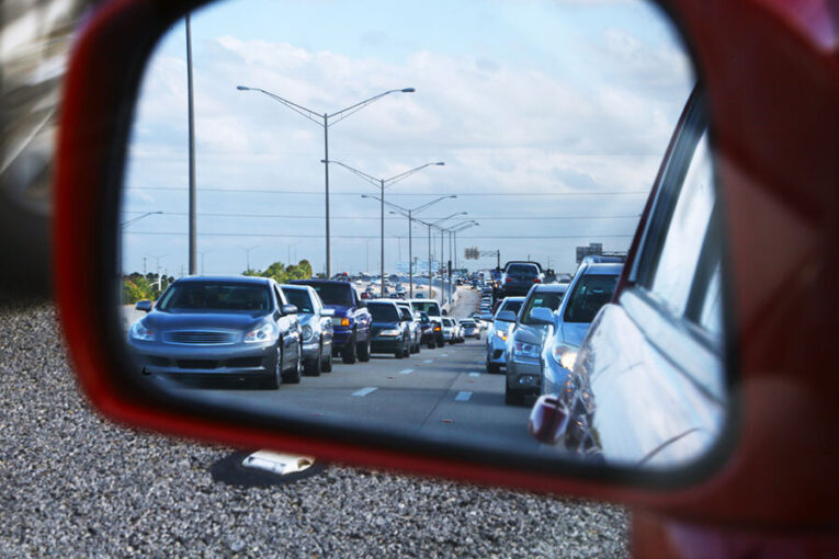 Traffic jam seen through a vehicle's side mirror, showing multiple lanes of closely packed cars on a highway under a partly cloudy sky.