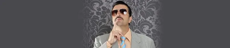 A man in a light gray suit, orange shirt, blue tie, and sunglasses stands against a patterned dark background, holding his finger to his lips.