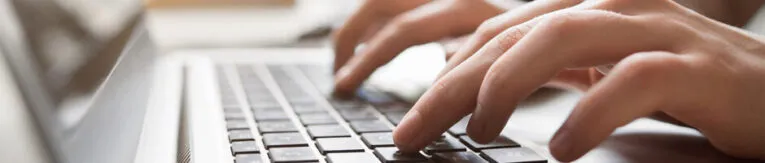 Close-up of a person typing on a laptop keyboard. The photo focuses on the person's hands and the keys. The laptop screen is not visible. The background is blurred.