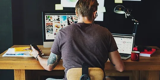 A person with a tattoo on their left arm works at a desk with two computer screens showcasing photos for your essentials website (updated 2024), a calendar on a laptop, a smartphone in hand, and a red mug nearby.
