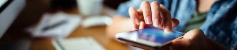 A person using a smartphone, with a keyboard and computer screen visible in the background, skillfully tracking mobile phone clicks.