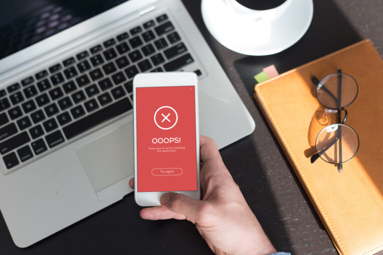 A person holds a smartphone displaying an error message, "OOOPS! There was an error making the request. Try again," in front of a laptop. A cup, glasses, and notebook are on the table—an all-too-common scene for those who need to learn about the 3 website errors you can fix yourself.