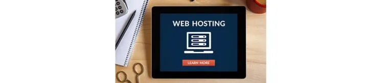 A tablet displaying a web hosting advertisement with a "Learn More" button, surrounded by a notebook, a pen, a pair of scissors, and a drink on a wooden surface.