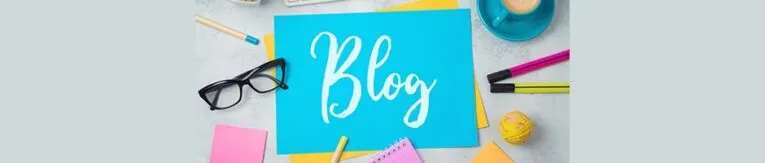 A blue paper with the word "Blog" written on it in white, surrounded by stationery items including glasses, markers, a cup, a ball, and sticky notes on a grey surface.