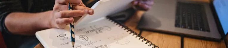 A person holds a pencil and a notepad with sketches and notes, next to an open laptop on a wooden desk.