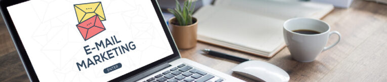 A laptop on a desk displays an "E-Mail Marketing" screen. Nearby are a potted plant, a notebook, a coffee cup, and a computer mouse.