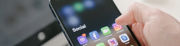 Close-up of a smartphone screen displaying a folder labeled "Social" with app icons for Facebook, Instagram, WhatsApp, and others. A hand is about to tap on the Instagram icon.