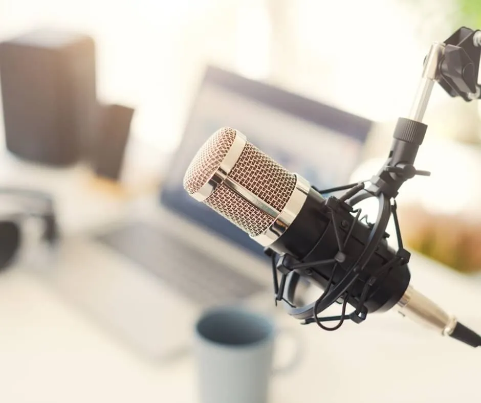 Close-up of a professional microphone on a boom stand in front of a blurred, open laptop and a coffee mug.