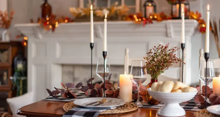 A dining table set for a meal, featuring candles, a centerpiece of autumn leaves, bread in a bowl, and plates with cutlery on woven placemats. A decorated mantel with seasonal items is in the background.