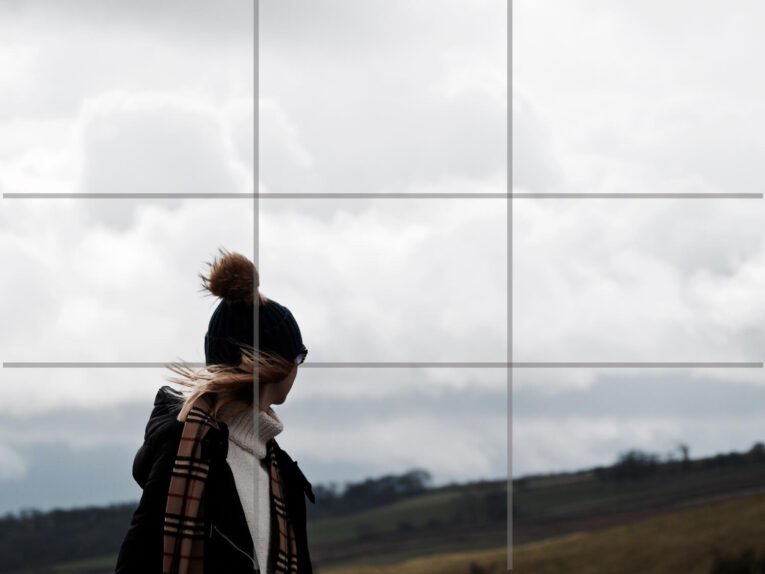 Person wearing a dark jacket and a beanie with a pom-pom stands outdoors looking at a cloudy sky.