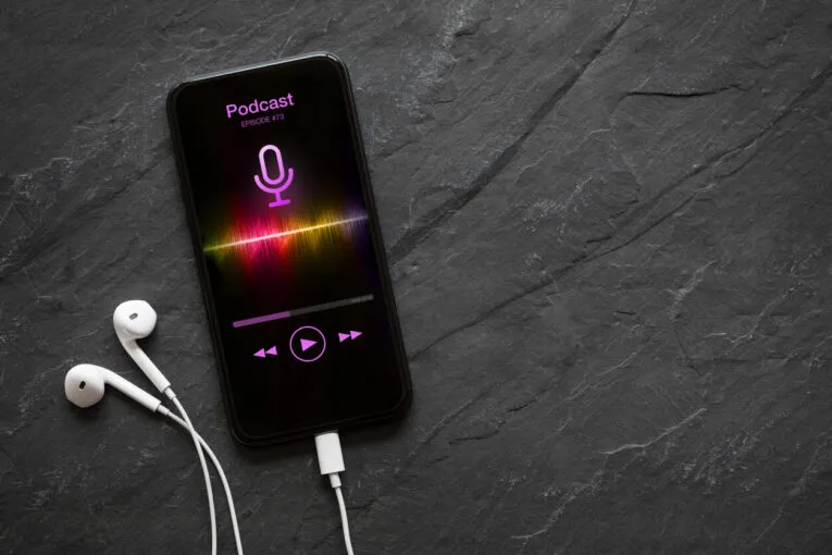 Smartphone on a dark gray surface displaying a podcast episode screen with earphones plugged in.