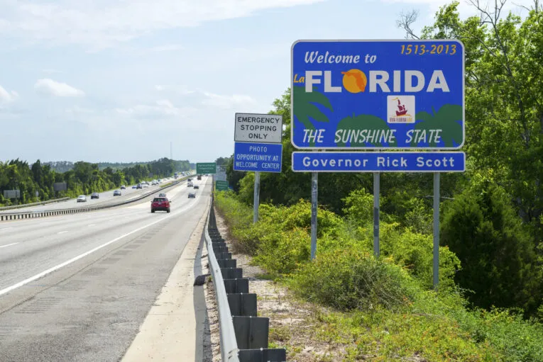 A sign reading "Welcome to Florida, The Sunshine State" stands beside a highway, with "Governor Rick Scott" and "1513-2013" mentioned. Several cars speed by on the greenery-lined road. Nearby, a smaller sign advises: “Florida Lawyers: Update Your Website Content to Reflect Recent Tort Reform Legislation.”