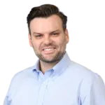 A man with dark hair and a beard is smiling while wearing a light blue button-up shirt against a white background.