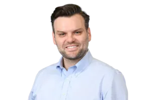 A man with dark hair and a beard is smiling while wearing a light blue button-up shirt against a white background.